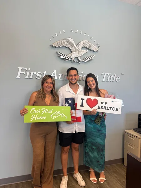 Three people in front of a First American Title wall sign holding Our First Home and I Love My Realtor signs
