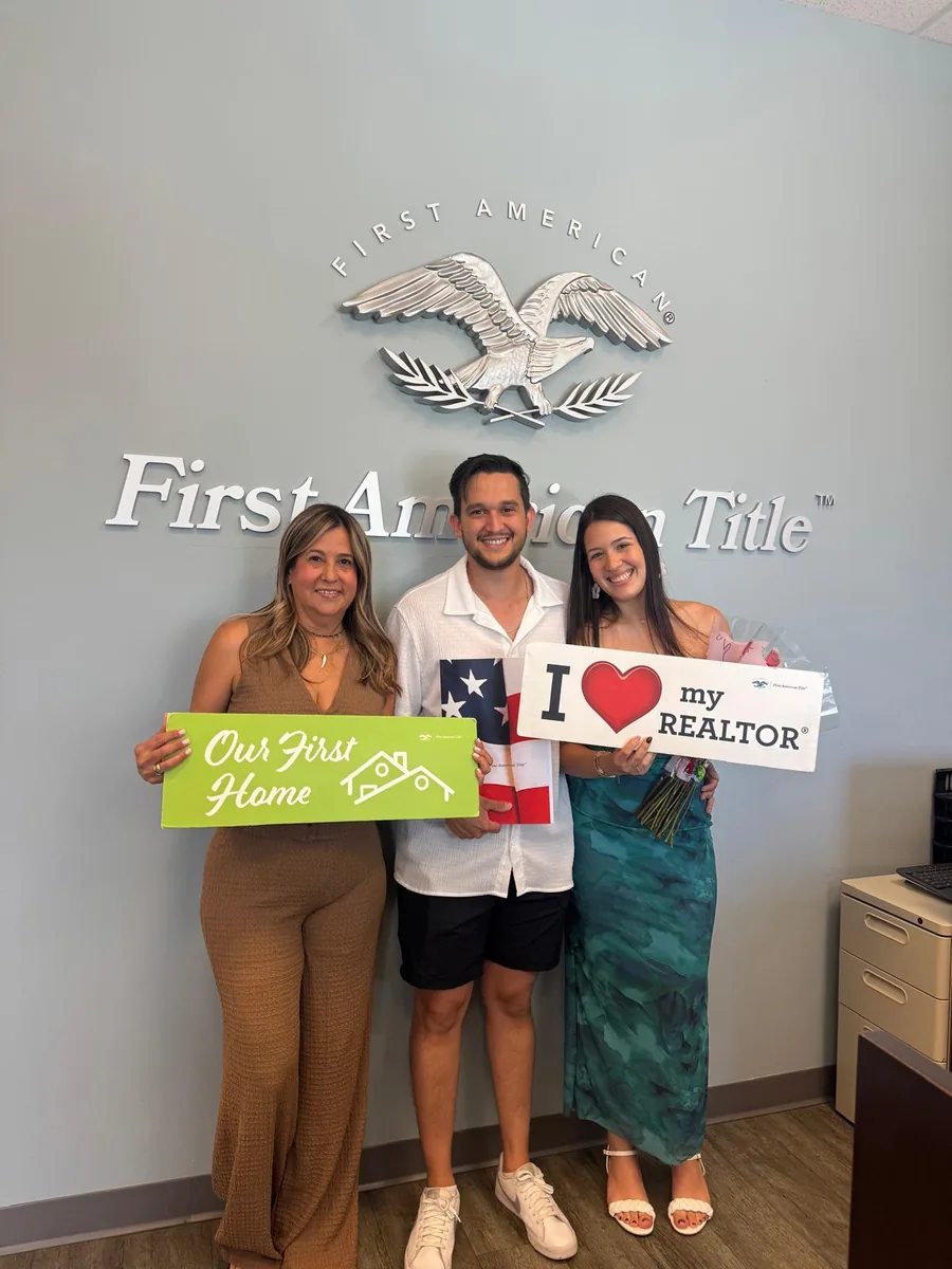 Three people in front of a First American Title wall sign holding Our First Home and I Love My Realtor signs