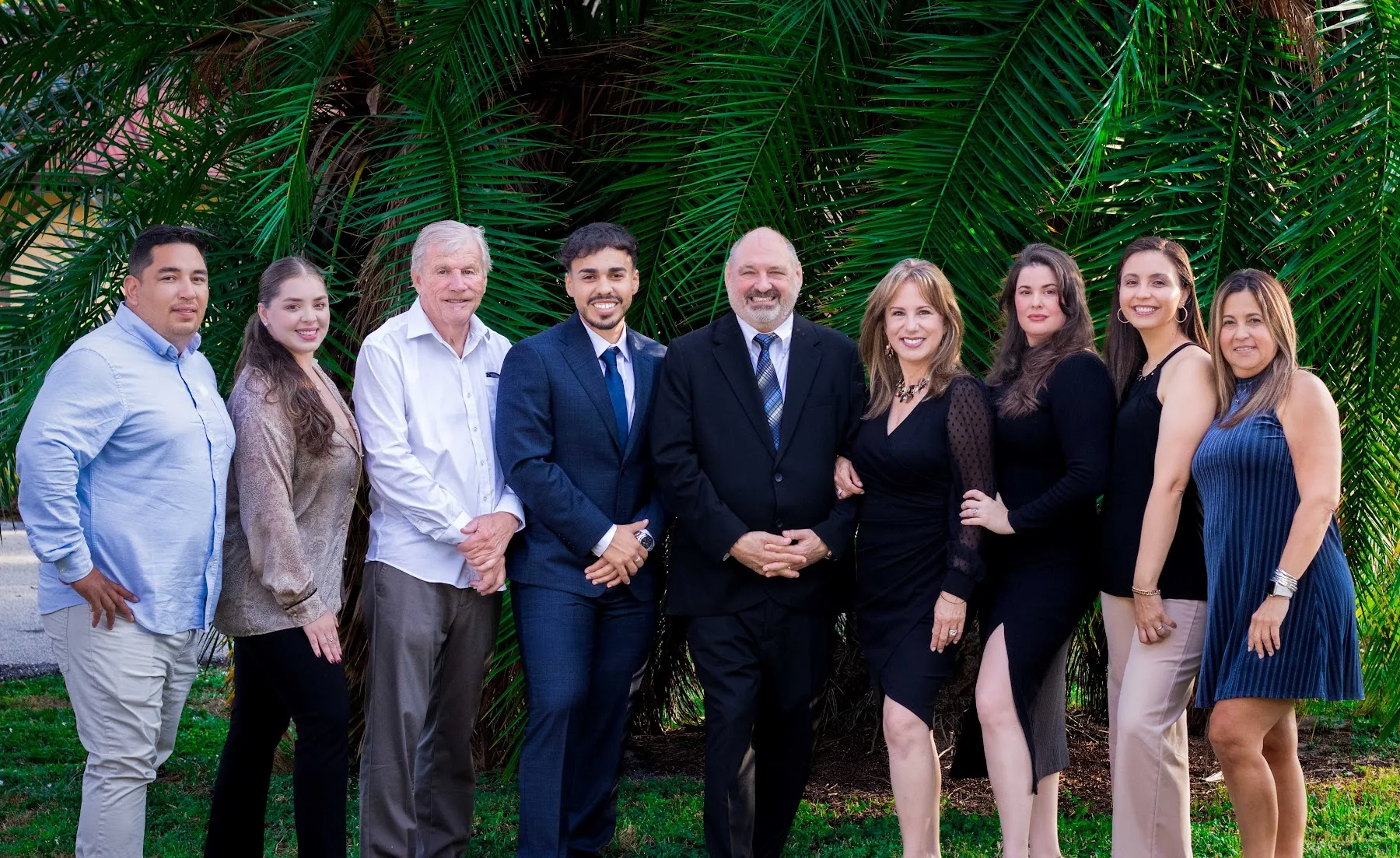 The Icon Premier Group team of nine agents and brokers standing together in business attire in front of tall palm fronds in Naples, Florida